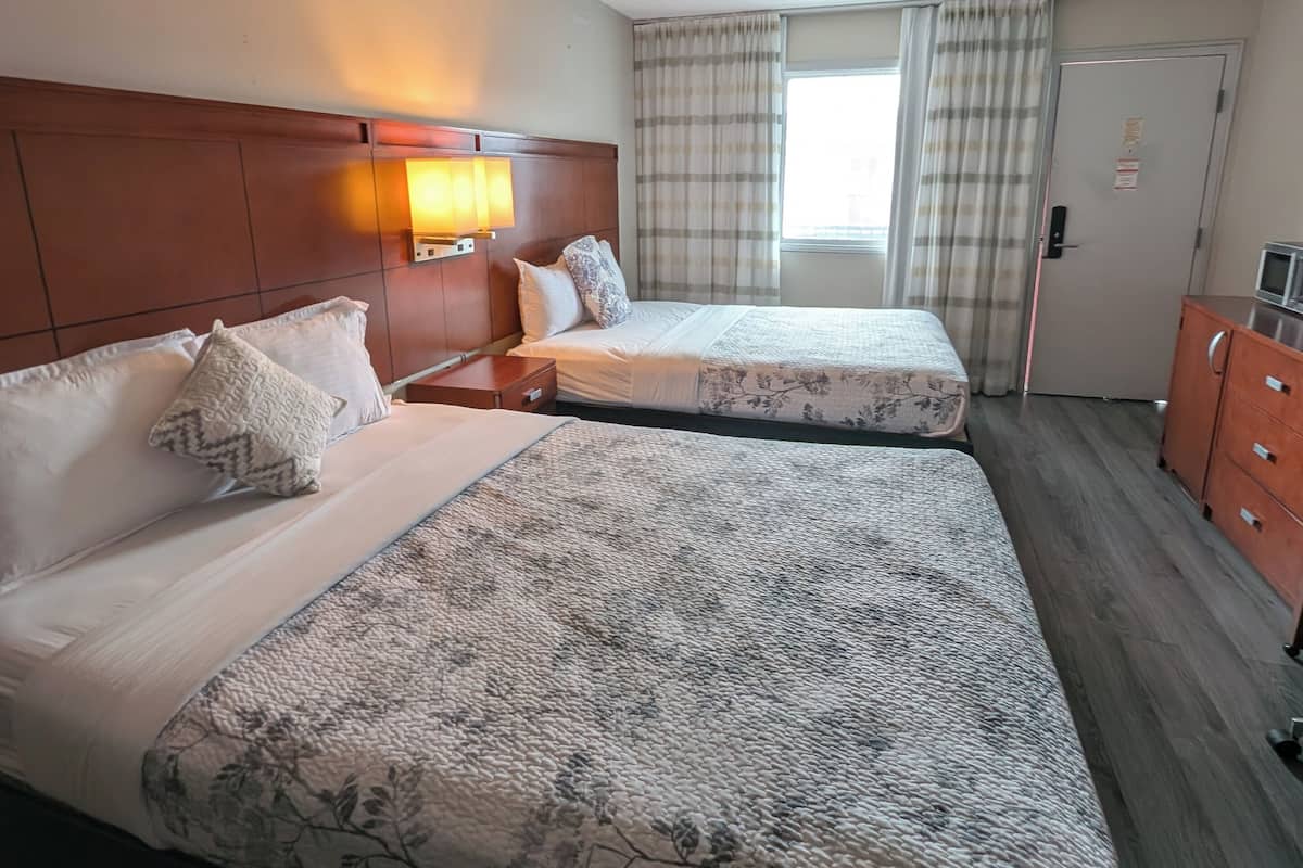 Ein Hotelzimmer mit zwei Betten, einem Holzkopfstück, einem Nachttisch mit Lampe, einem Schminktisch und einem Fenster mit Vorhängen.
