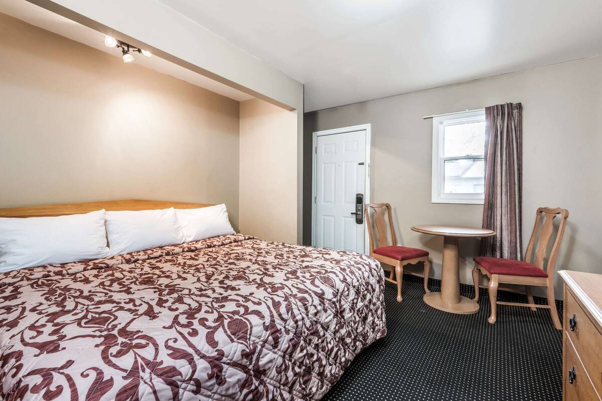A hotel room with a bed, a small table with chairs, a window with curtains, and a door.