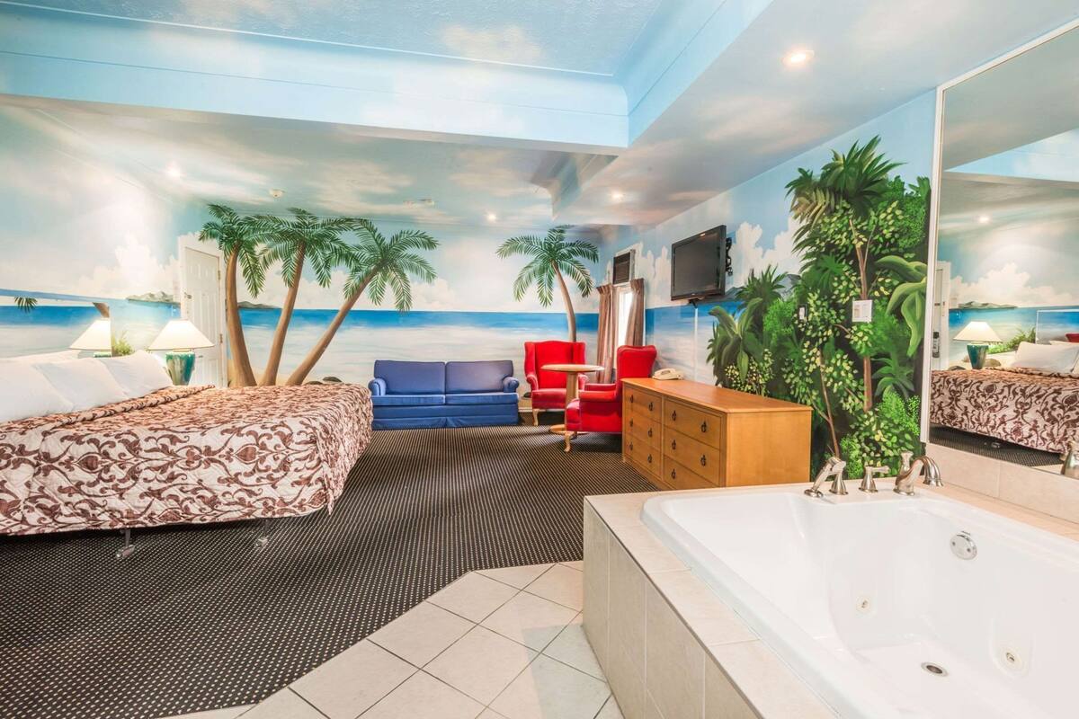 A hotel room with a bed, a sofa, a red chair, a wooden cabinet, a bathtub, and a large mirror.