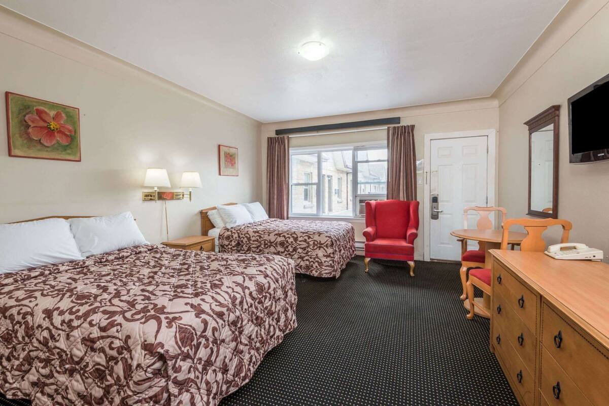A hotel room with two beds, a red armchair, a wooden dresser, a desk with a telephone, and a window with curtains.