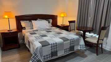 Ein ordentlich bezogenes Bett mit kariertem Überwurf, zwei Nachttischlampen, ein Holznachttisch, ein Stuhl und ein kleiner runder Tisch.