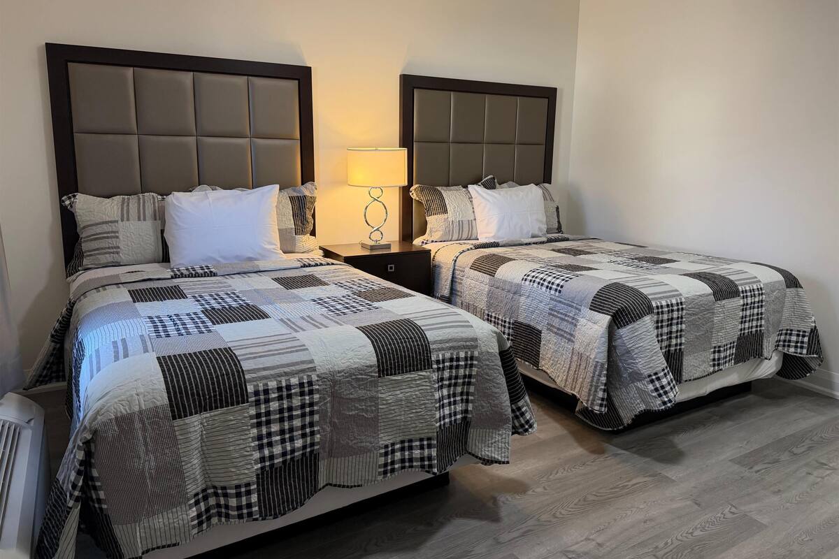 A hotel room with two beds, each with a quilted headboard, a bedside table with a lamp, and a wooden floor.