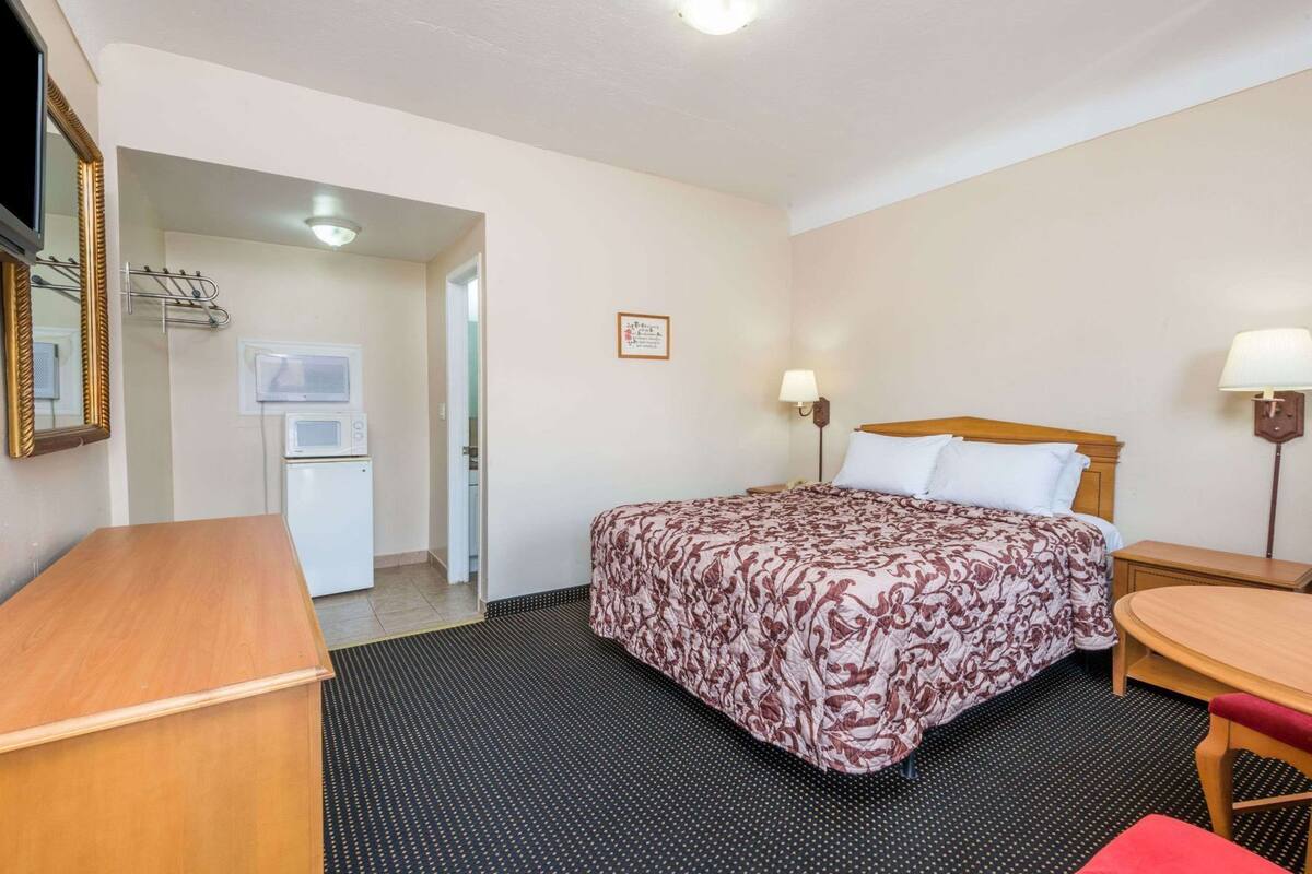 A hotel room with a bed, a desk, a chair, a lamp, a mirror, and a door leading to a bathroom.