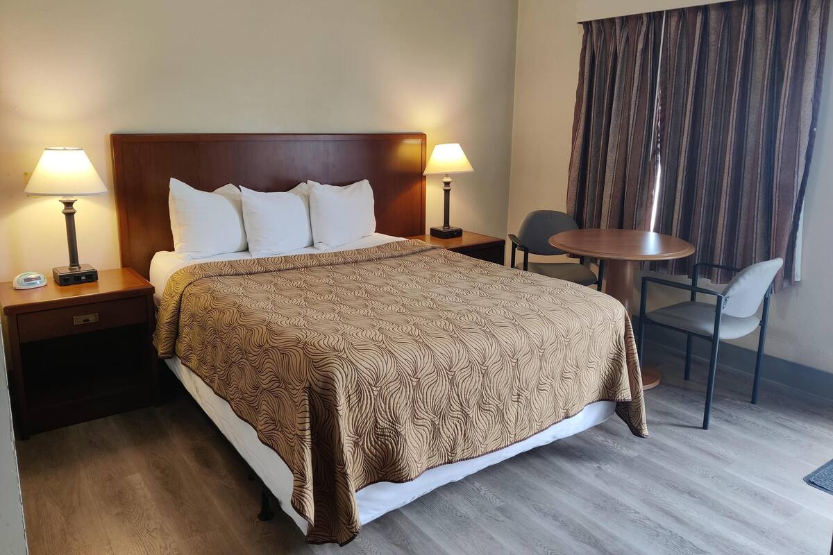A hotel room with a large bed, two bedside tables with lamps, a small round table with two chairs, and a window with curtains.