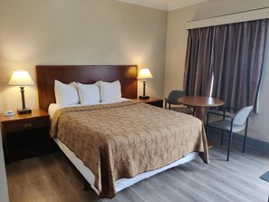 A hotel room with a large bed, two bedside tables with lamps, a small round table with two chairs, and a window with curtains.