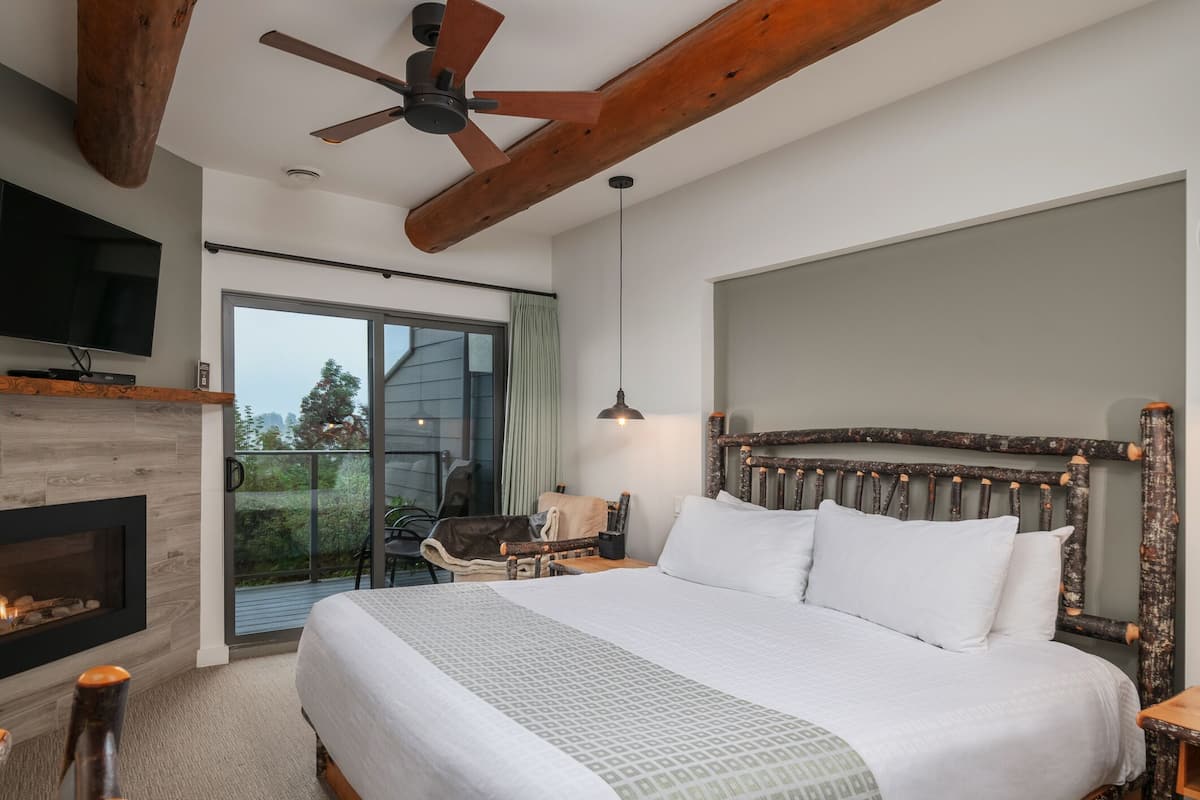 A bedroom with a large bed, a wooden headboard, a ceiling fan, a fireplace, a TV, and a balcony with a chair and a table.