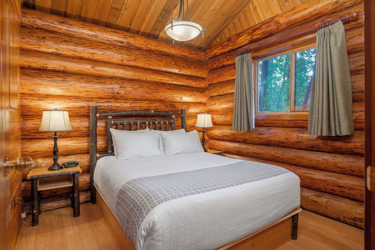 A log cabin bedroom with a wooden bed, two bedside lamps, and a window with curtains.