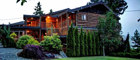 A log cabin with a balcony, surrounded by a well-maintained garden and tall trees.