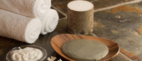 A spa setting with rolled white towels, a candle, a wooden bowl with a green substance, and a small bowl of cream.