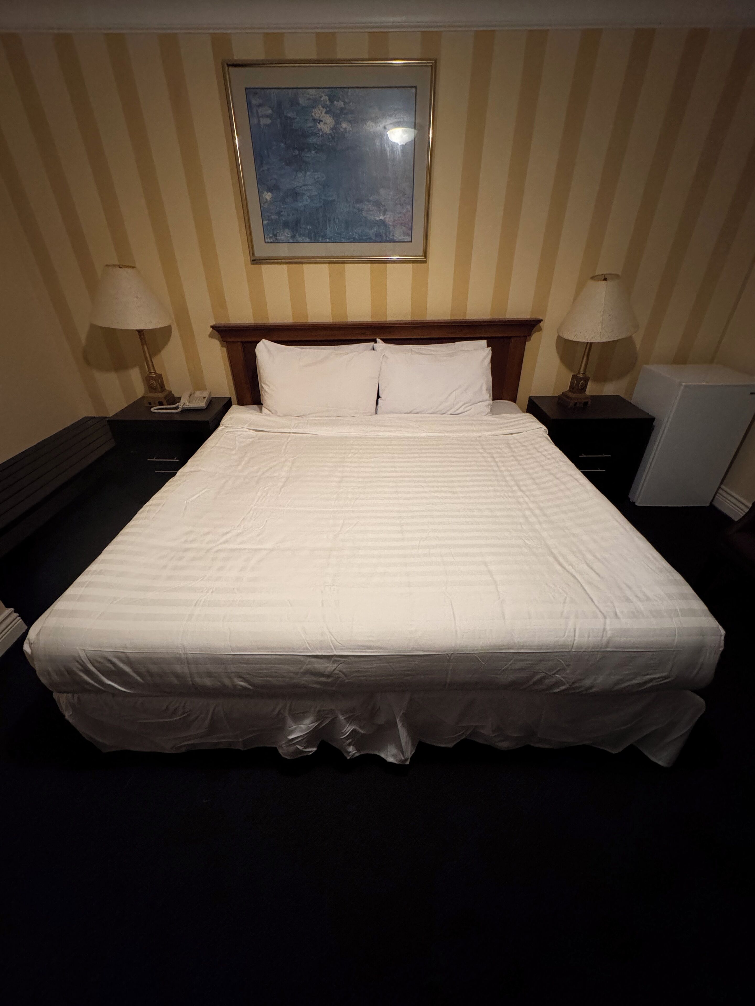 A neatly made bed in a hotel room with striped wallpaper, two bedside tables with lamps, and a framed picture on the wall.