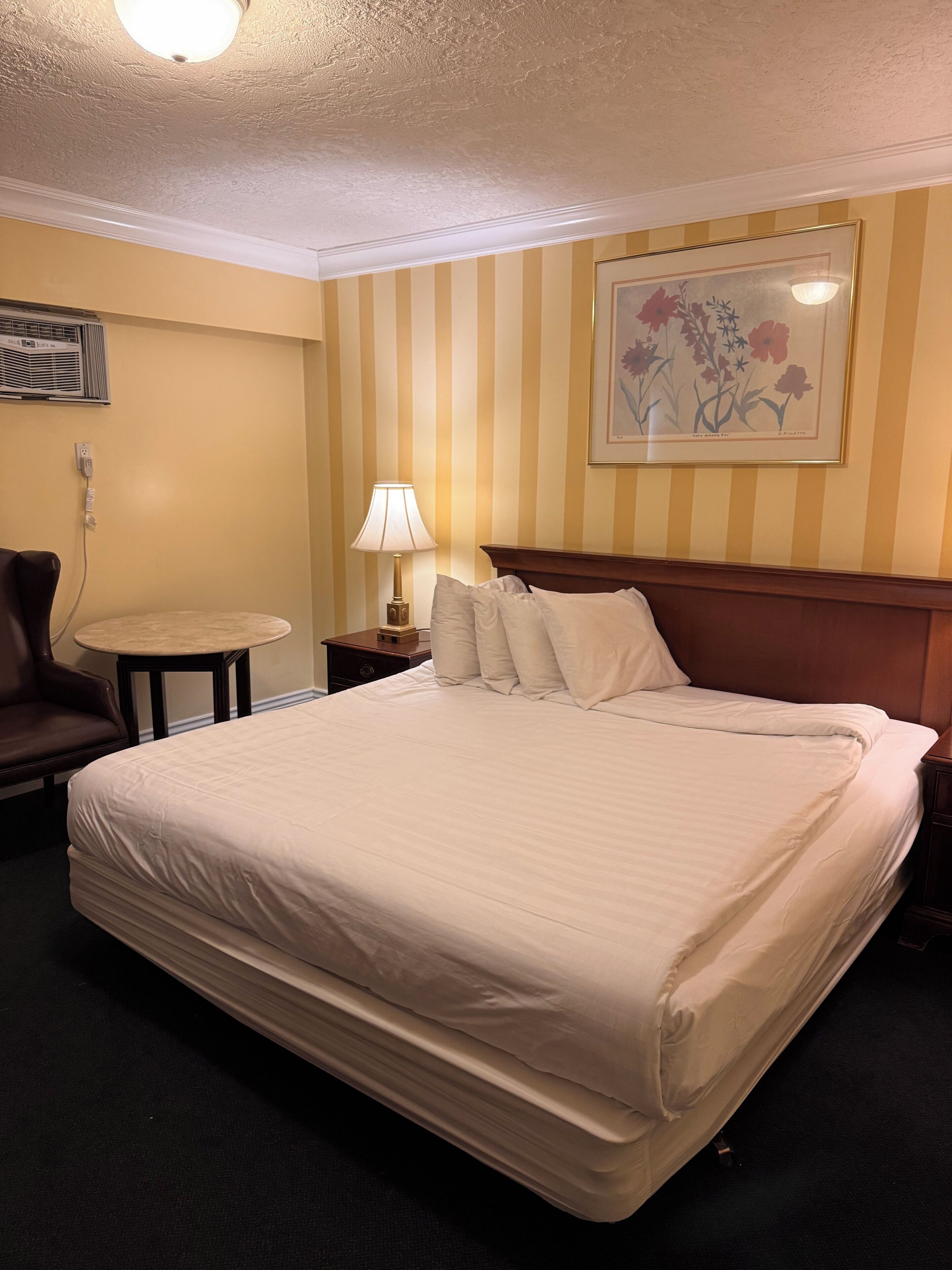 A hotel room with a large bed, a chair, a small table, a lamp, and a painting on the wall.