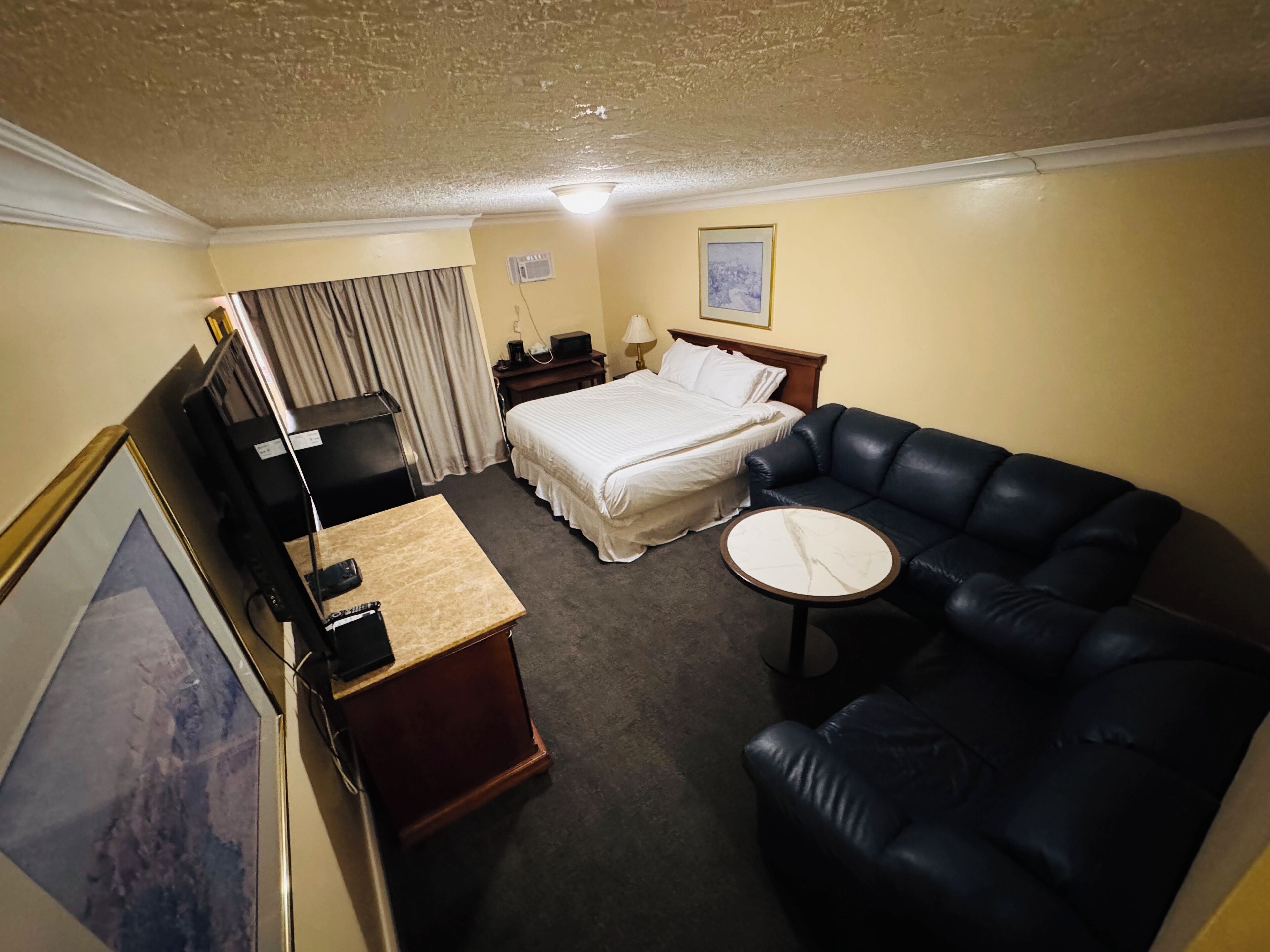 Hotelkamer met een bed, een zwarte leren bank, een klein rond tafeltje, een bureau met een computer, een televisie en een lijst aan de muur.