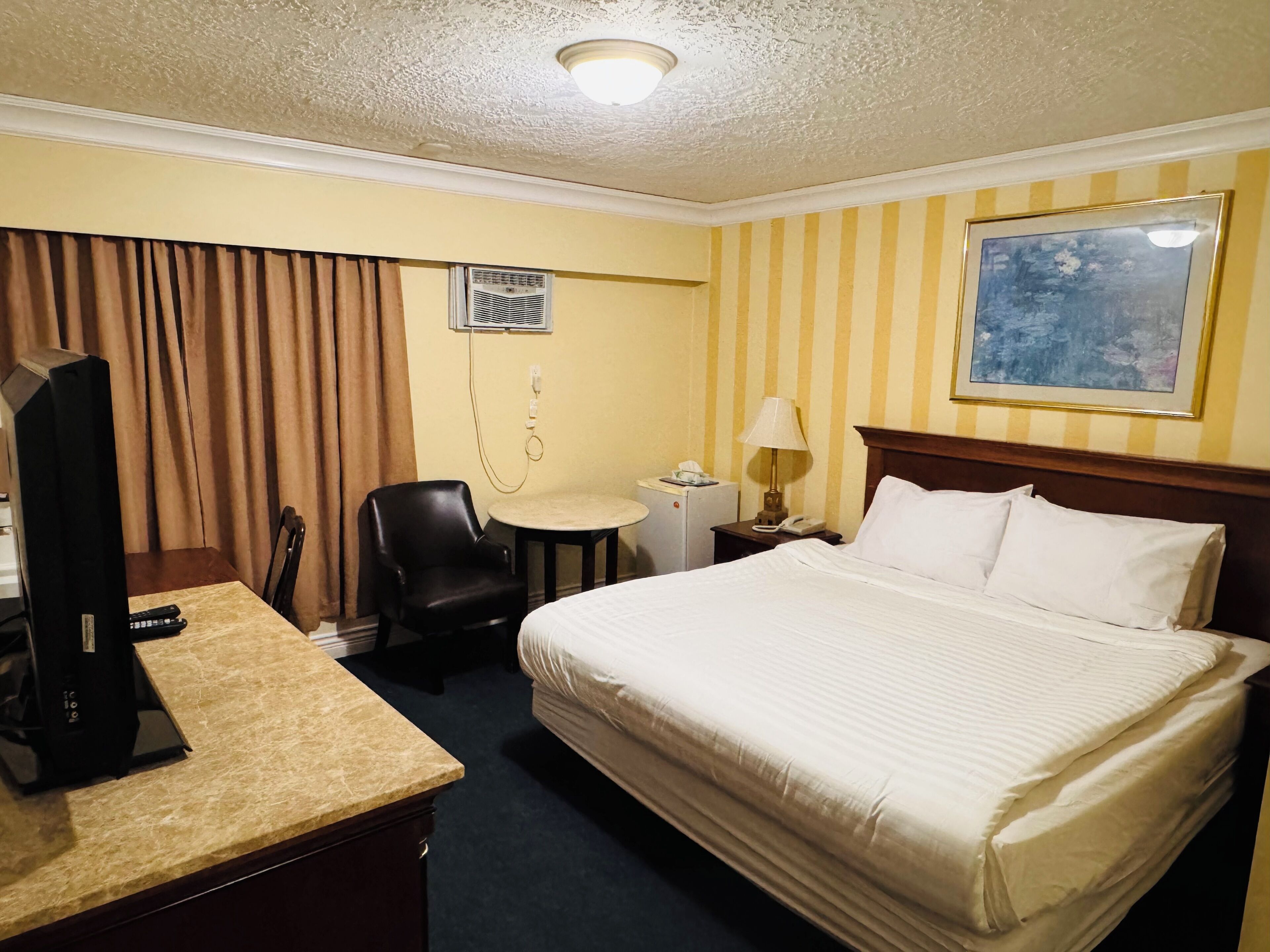 Une chambre d’hôtel avec un lit, un bureau avec un ordinateur, une chaise, une petite table, une lampe et un tableau au mur.
