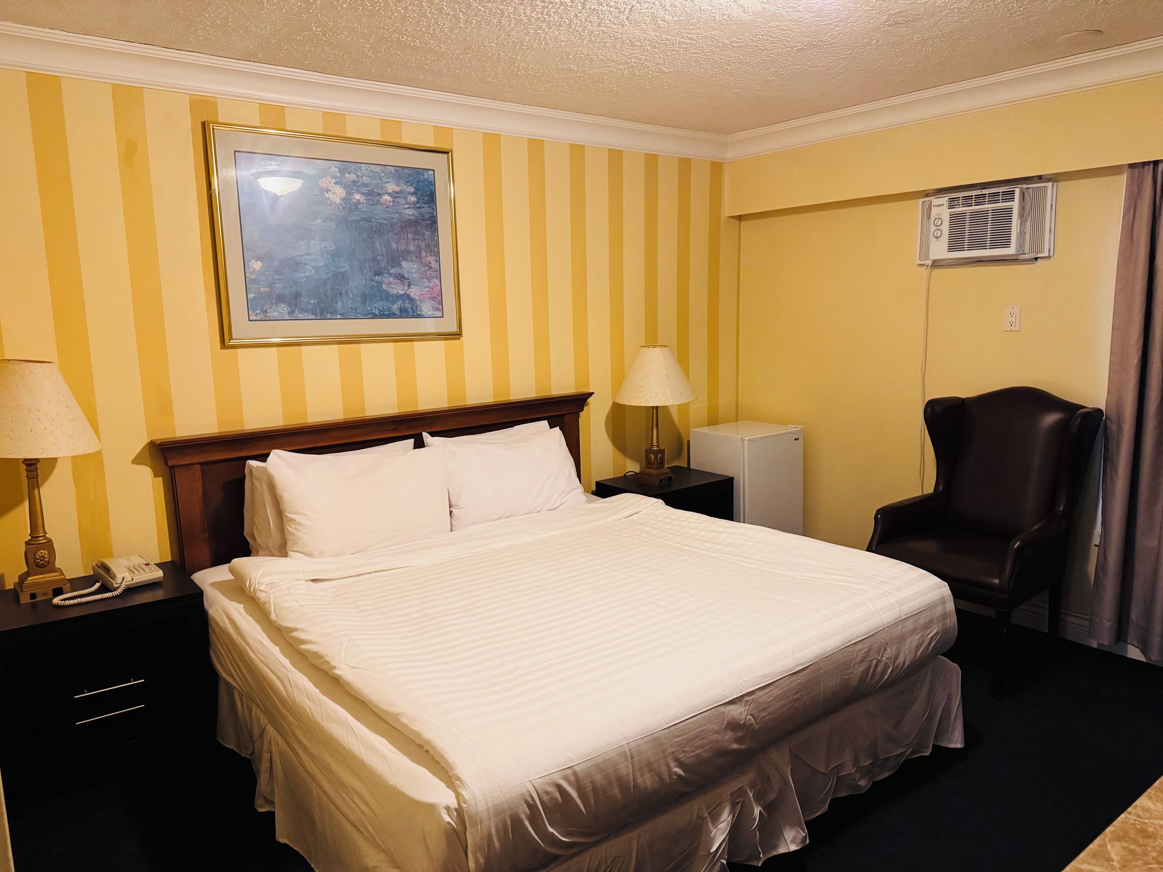 Habitación de hotel con una cama, dos lámparas de noche, un teléfono, un refrigerador blanco pequeño, una silla y un cuadro en la pared.