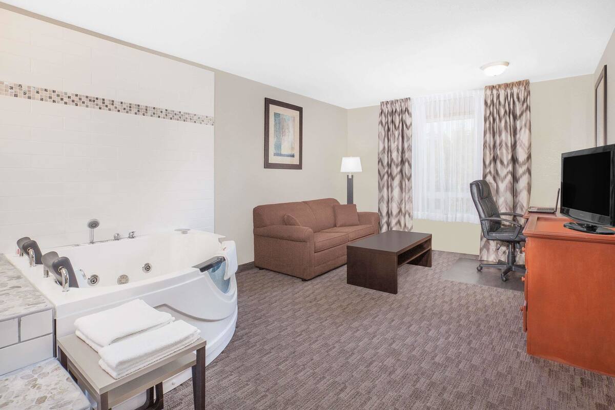 A hotel room with a large bathtub, a brown sofa, a desk with a television, and a chair.