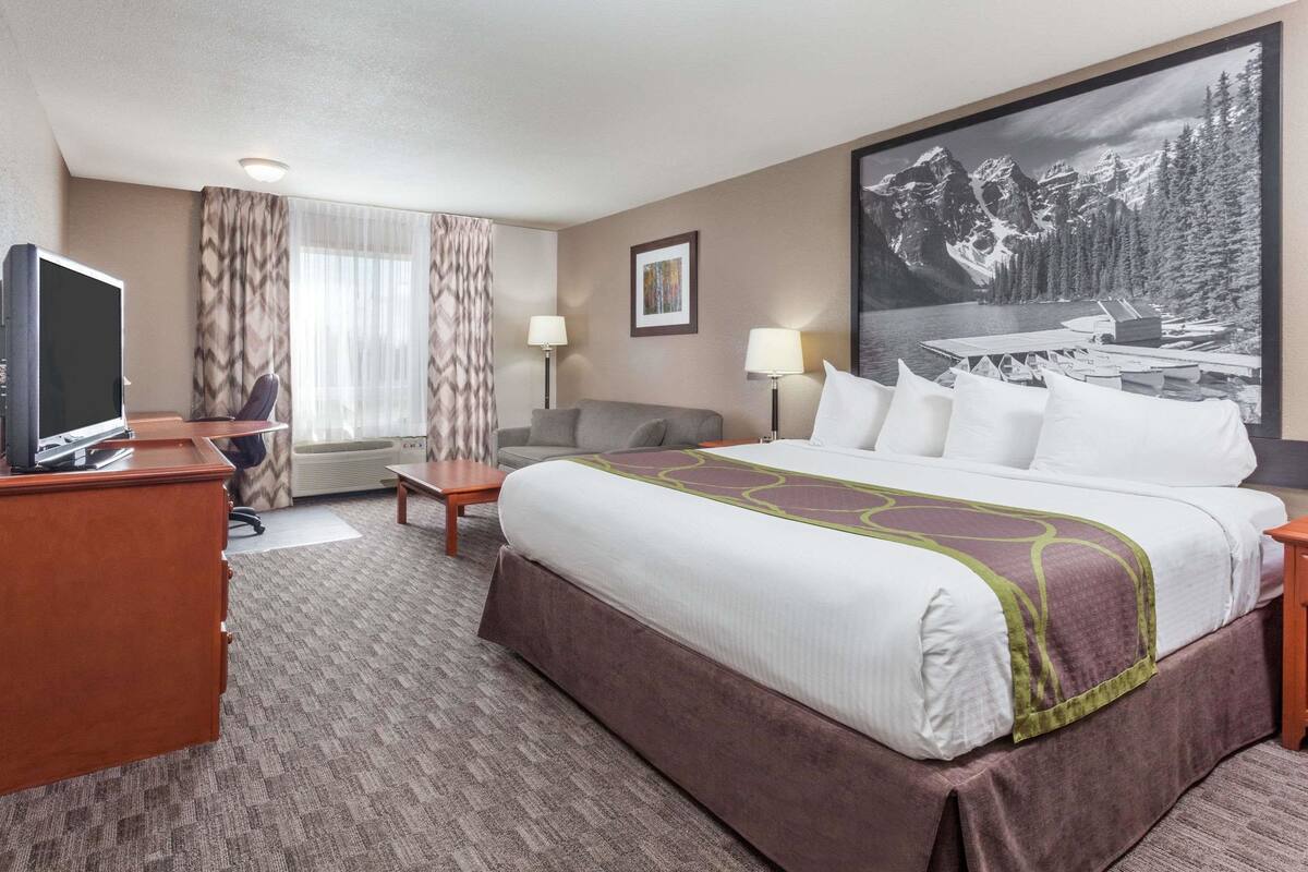 A hotel room with a large bed, a desk with a computer, a chair, a couch, and a large window with curtains.