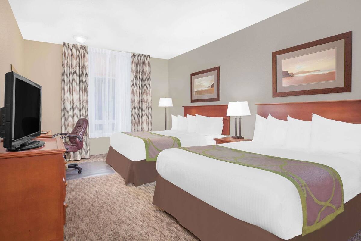 A hotel room with two beds, a desk with a TV, a chair, a desk lamp, and two framed pictures on the wall.