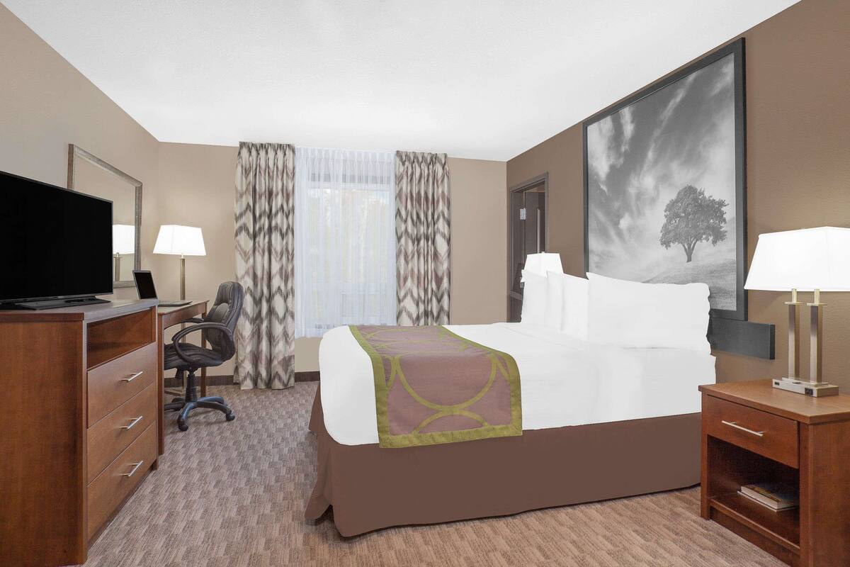 A hotel room with a bed, a desk with a computer, a chair, a TV, a lamp, and a painting on the wall.