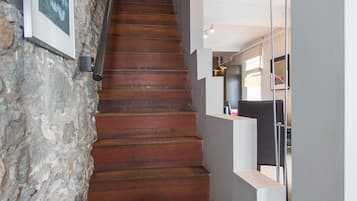 Un escalier en bois mène à un palier, lequel est bordé d’un côté par un mur de pierre et de l’autre par un mur blanc doté d’étagères intégrées.