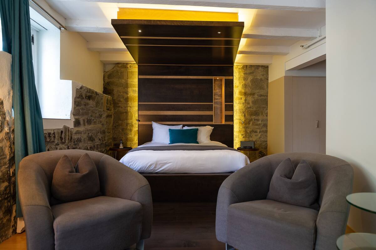 A modern hotel room with a stone accent wall, a large bed with white bedding, two grey armchairs, and a glass side table.