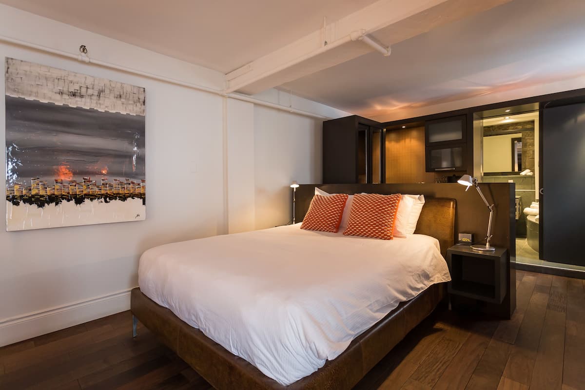A modern bedroom with a large bed, a nightstand with a lamp, and a wall-mounted artwork.