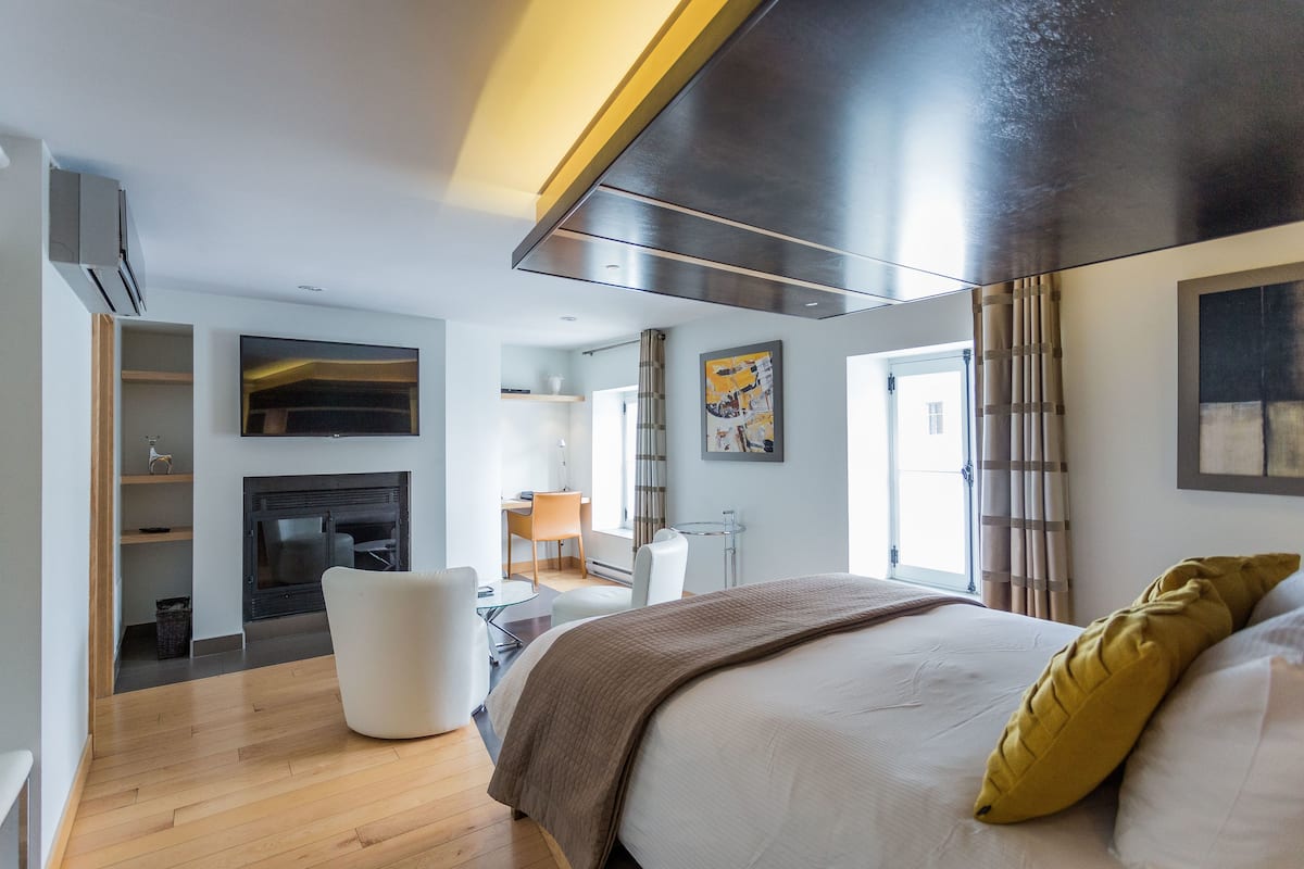 A modern hotel room with a large bed, a fireplace, a desk, and a view of the city.