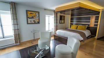 A modern bedroom with a bed, two white chairs, a glass table, a painting, and a large window.