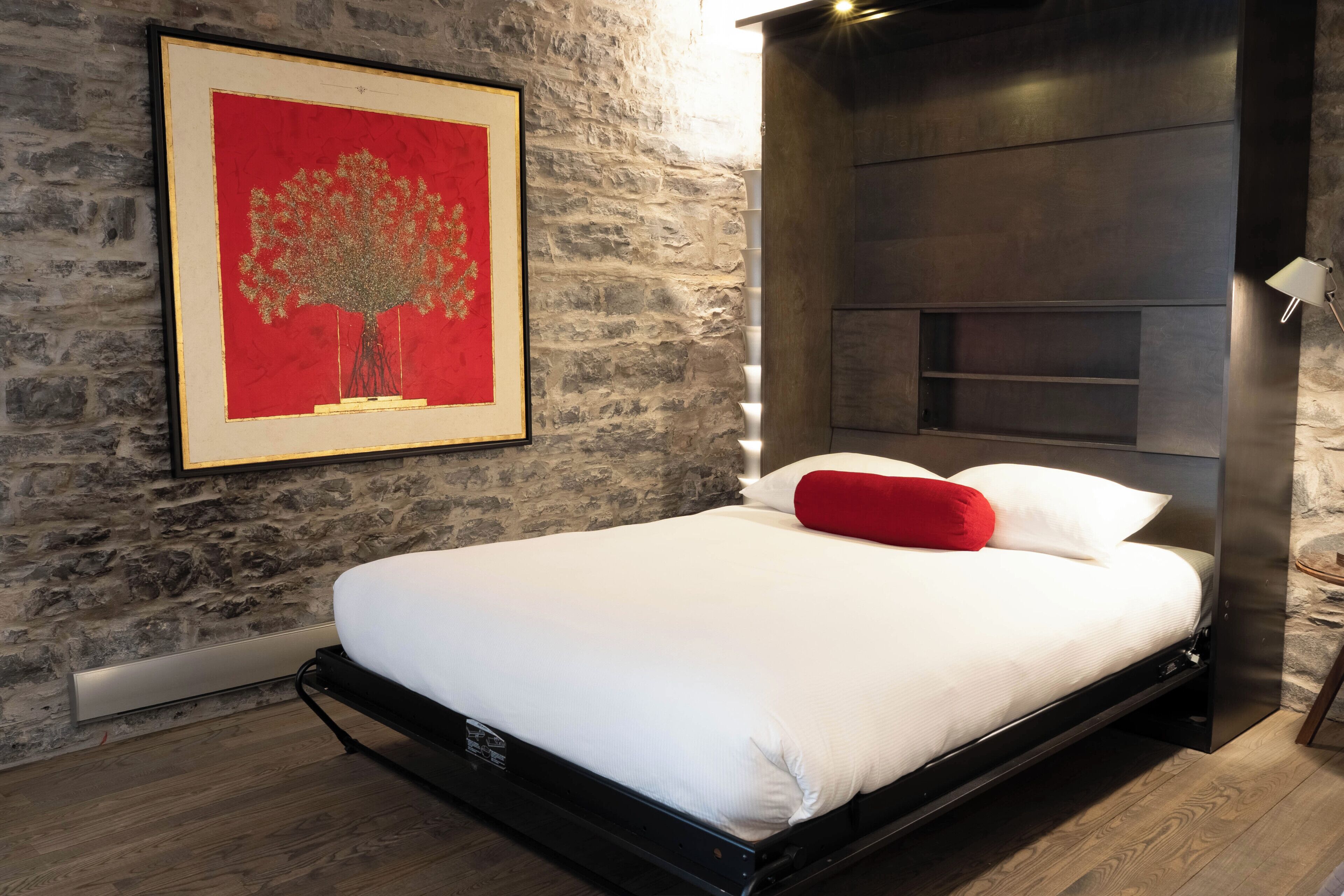Un dormitorio moderno con pared de piedra, una obra de arte enmarcada, una cama con ropa blanca y almohadas rojas, y cabecero integrado.