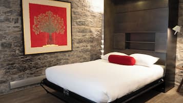 Un dormitorio moderno con pared de piedra, una obra de arte enmarcada, una cama con ropa blanca y almohadas rojas, y cabecero integrado.