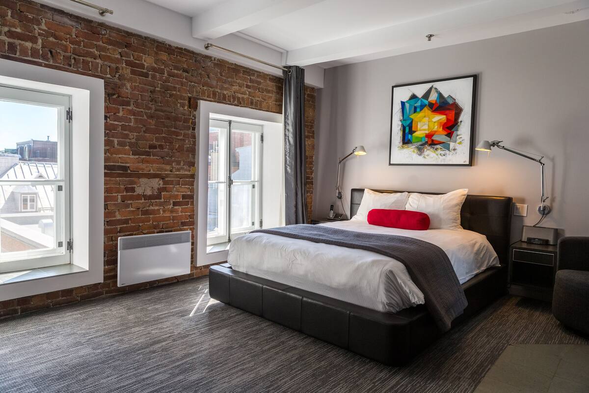 A modern bedroom with a brick wall, a large bed with a gray and white duvet, a red pillow, and a colorful abstract painting on the wall.