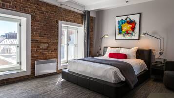 A modern bedroom with a brick wall, a large bed with a gray and white duvet, a red pillow, and a colorful abstract painting on the wall.