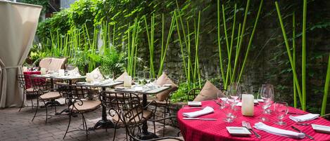 Un área de comedor al aire libre con mesas dispuestas para dos, rodeada de abundante follaje y plantas de bambú.