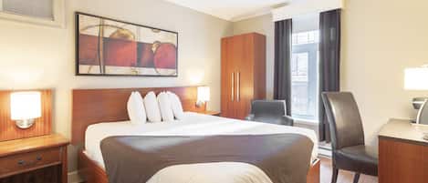A hotel room with a large bed, wooden headboard, two bedside tables with lamps, a desk with a chair, a large window with curtains, and a painting on the wall.