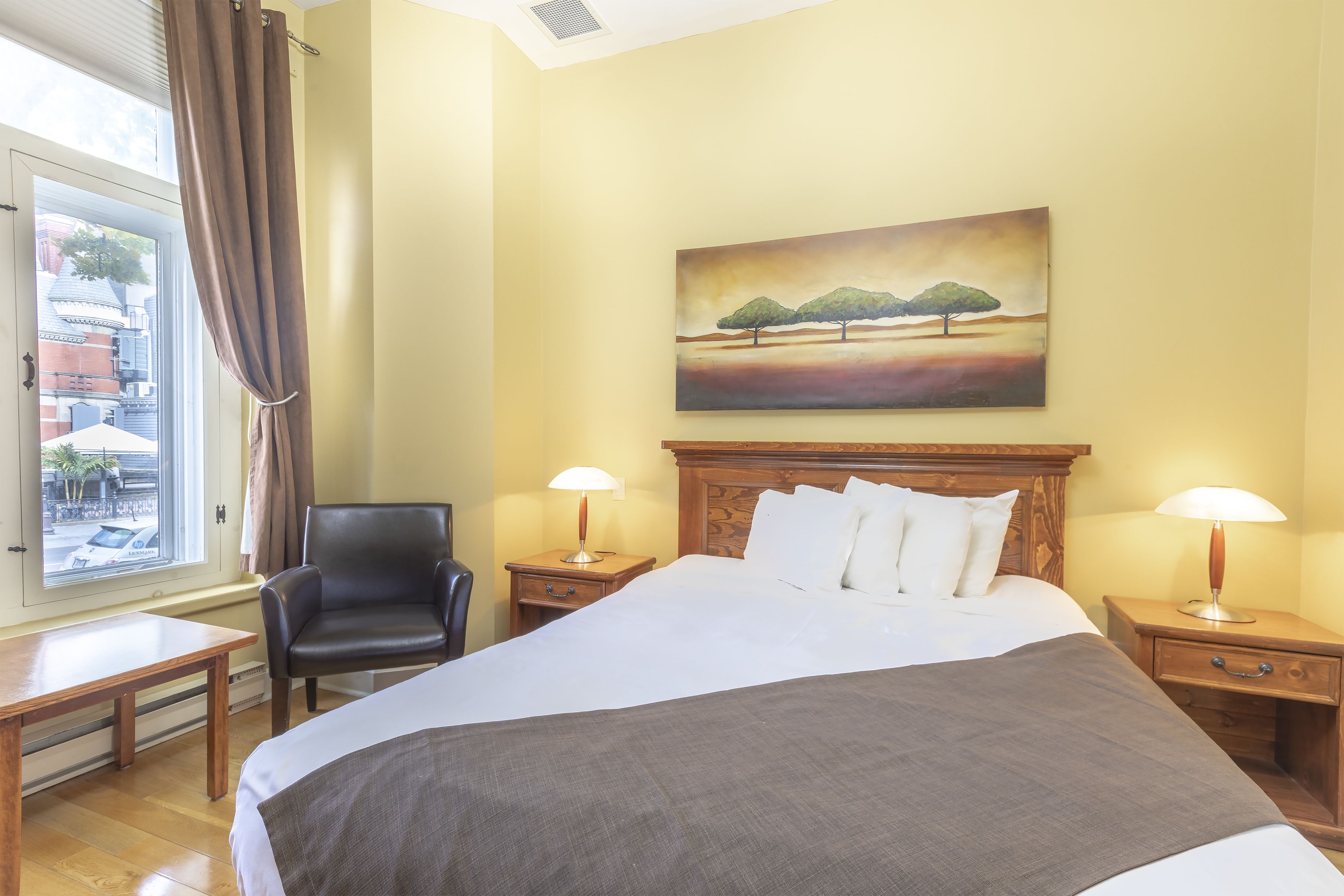 Habitación de hotel con una cama, dos mesitas de noche con lámparas, una silla, una ventana con cortinas y un cuadro en la pared.