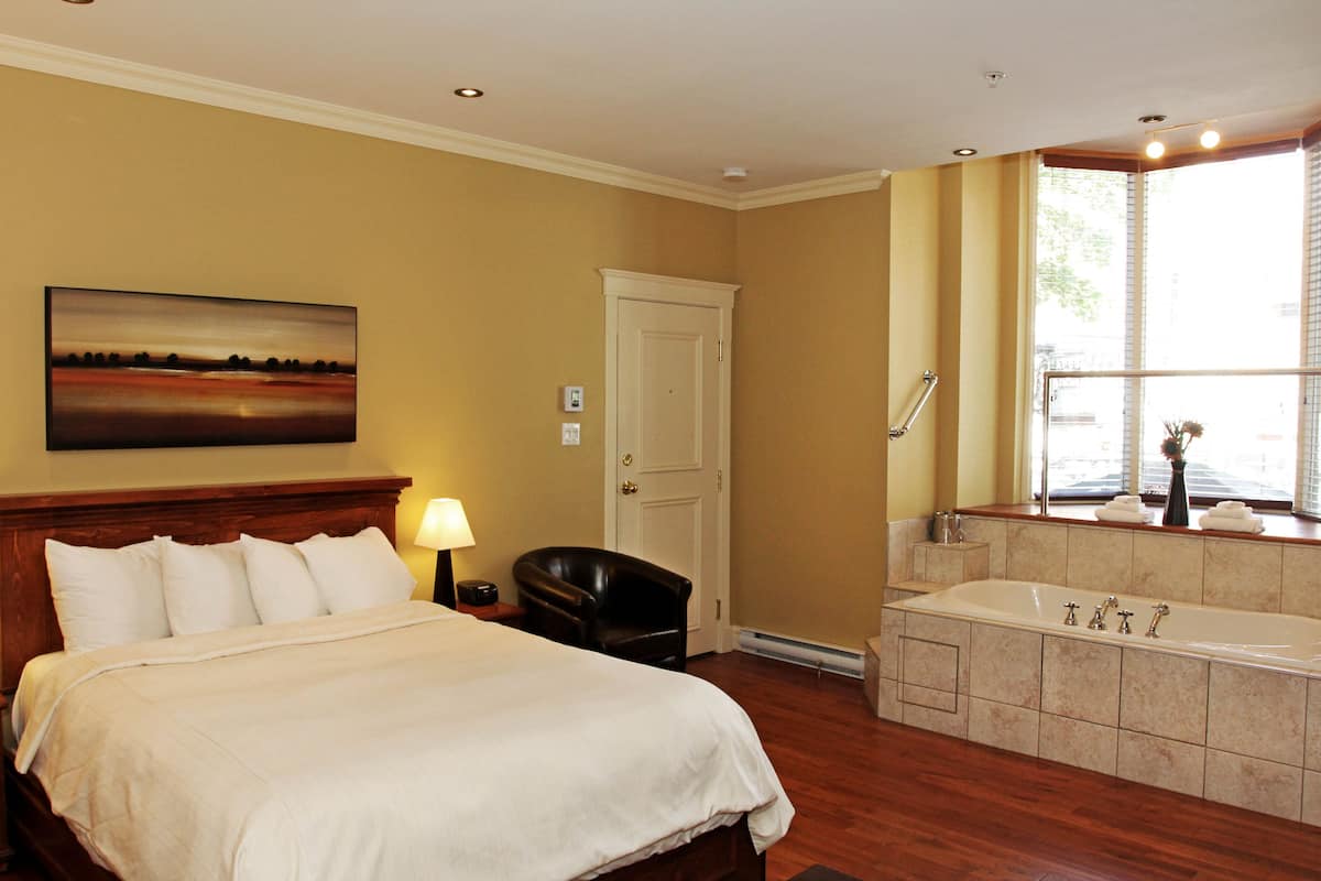 Habitación de hotel con una cama grande, una zona de estar con un sillón, un baño con bañera y un cuadro en la pared.