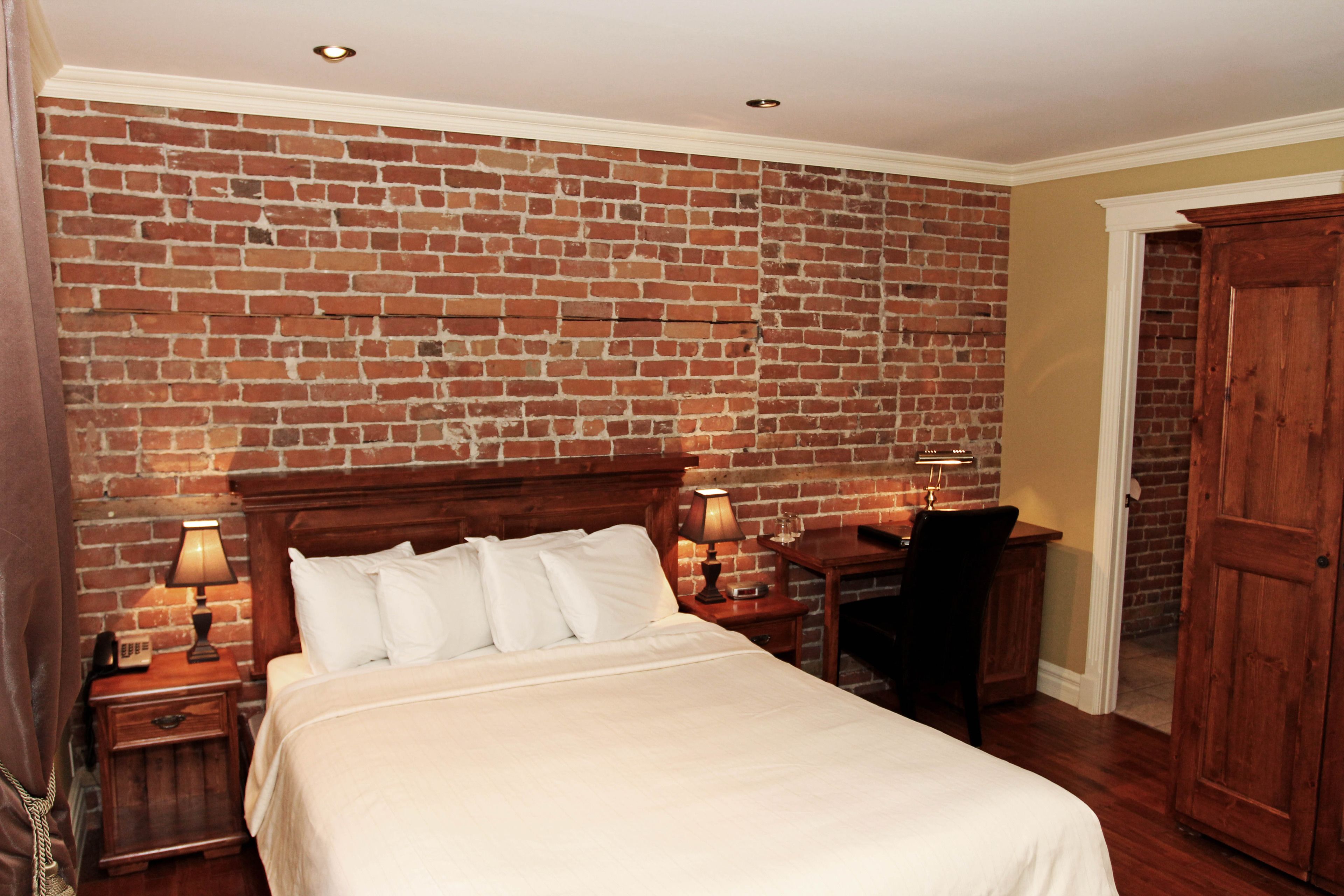 Ein Hotelzimmer mit Backsteinwand, einem hölzernen Bett, zwei Nachttischen mit Lampen, einem Schreibtisch und einem Kleiderschrank.