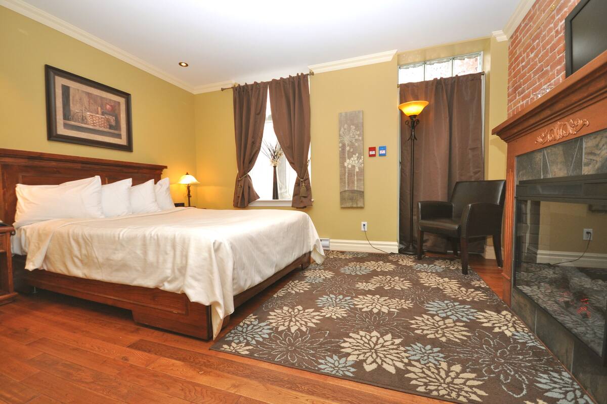 Una habitación de hotel con una cama de madera, una chimenea, un ventanal con cortinas y una alfombra estampada con motivos florales.