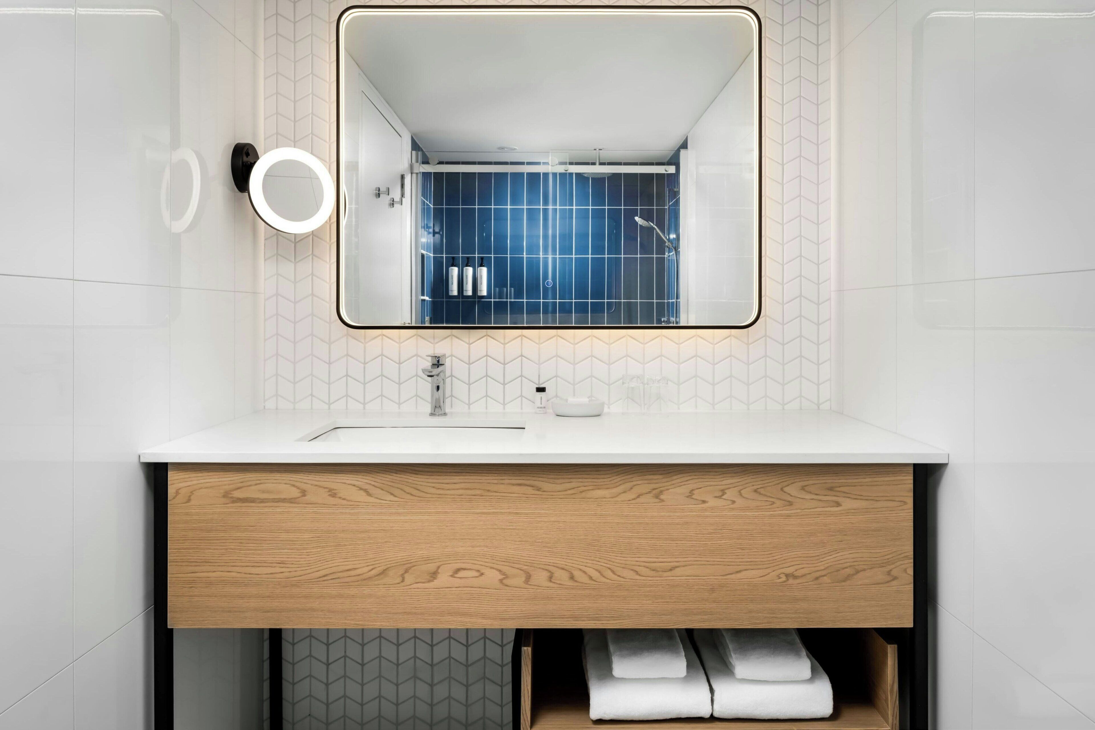 A modern bathroom with a white sink, wooden vanity, and a mirror with a lighted frame.