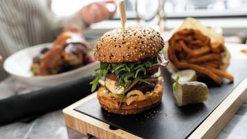 A burger with a sesame seed bun, lettuce, and a toothpick, served on a wooden board with fries and a pickle in the background.