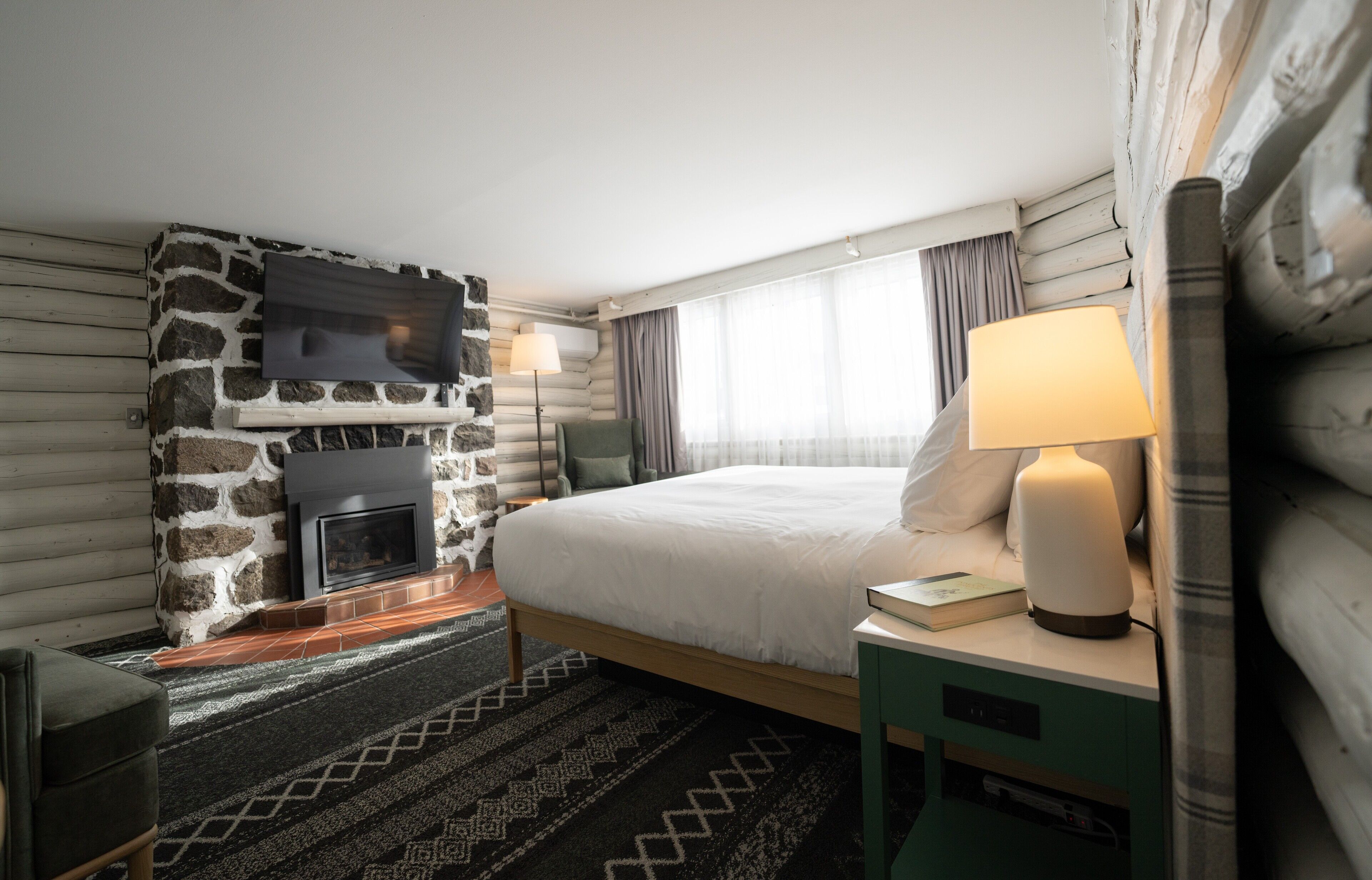 Hotellihuone, jossa on kivinen takka, valkoisilla lakanoilla varustettu sänky, yöpöytä lampun kanssa ja seinälle asennettu televisio.