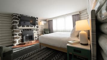 Hotellihuone, jossa on kivinen takka, valkoisilla lakanoilla varustettu sänky, yöpöytä lampun kanssa ja seinälle asennettu televisio.