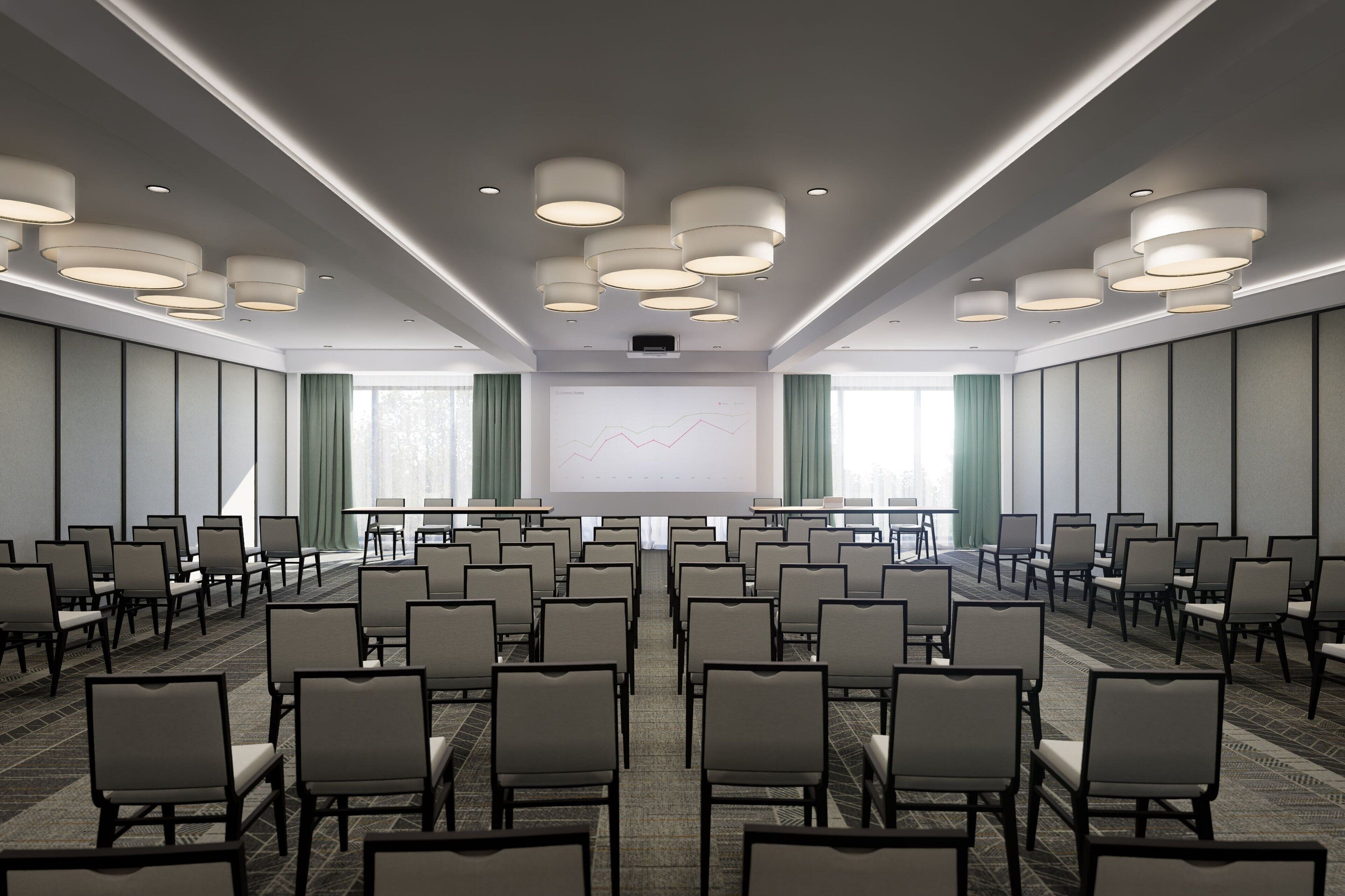 Un salón de conferencias con filas de sillas orientadas hacia una pantalla de proyección, luces empotradas en el techo y un amplio ventanal con cortinas.