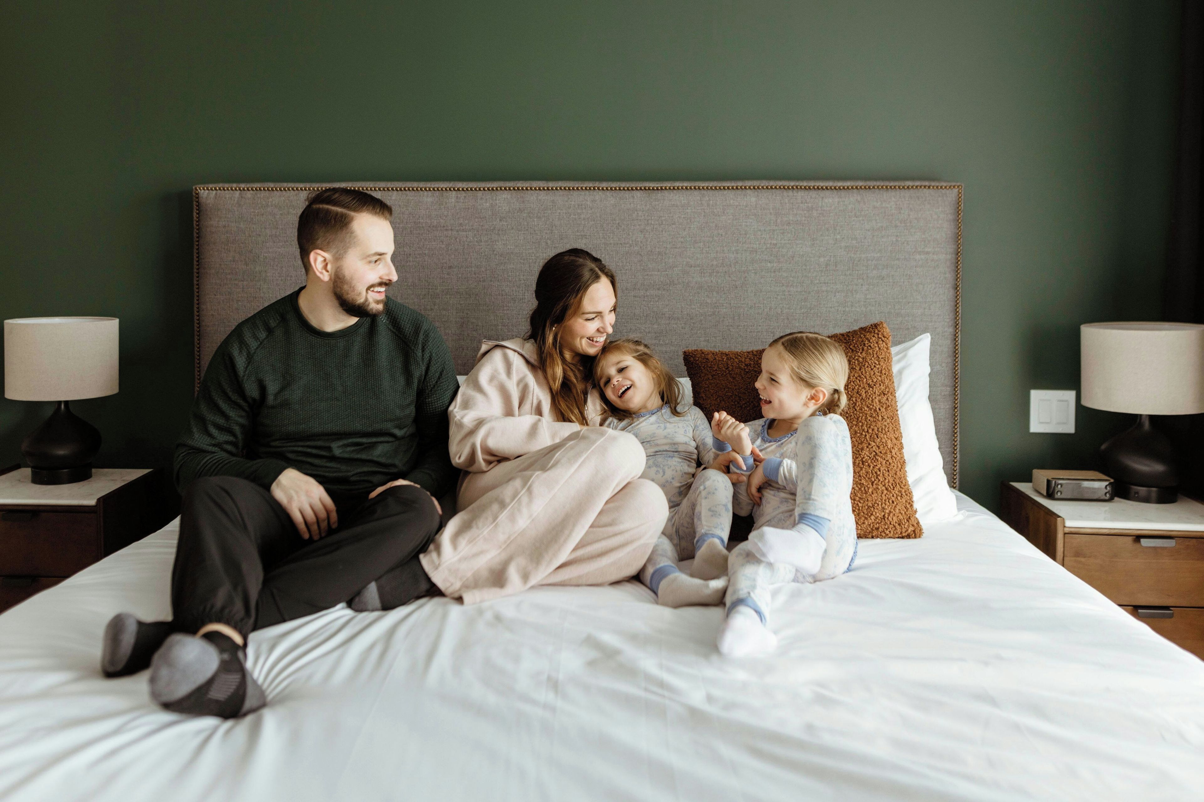 Una familia de cuatro sobre una cama con cabecera, dos mesitas de noche con lámparas y una pared verde.