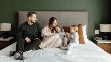 Una familia de cuatro sobre una cama con cabecera, dos mesitas de noche con lámparas y una pared verde.