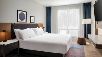 A hotel room with a large bed, a nightstand, a desk, a chair, a TV, and a window with curtains.