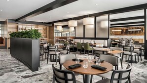 A modern restaurant with round tables, black chairs, and a central planter with green plants. - Mont Gabriel, Sainte-Adele, a Tribute Portfolio Resort (Sainte-Adele)