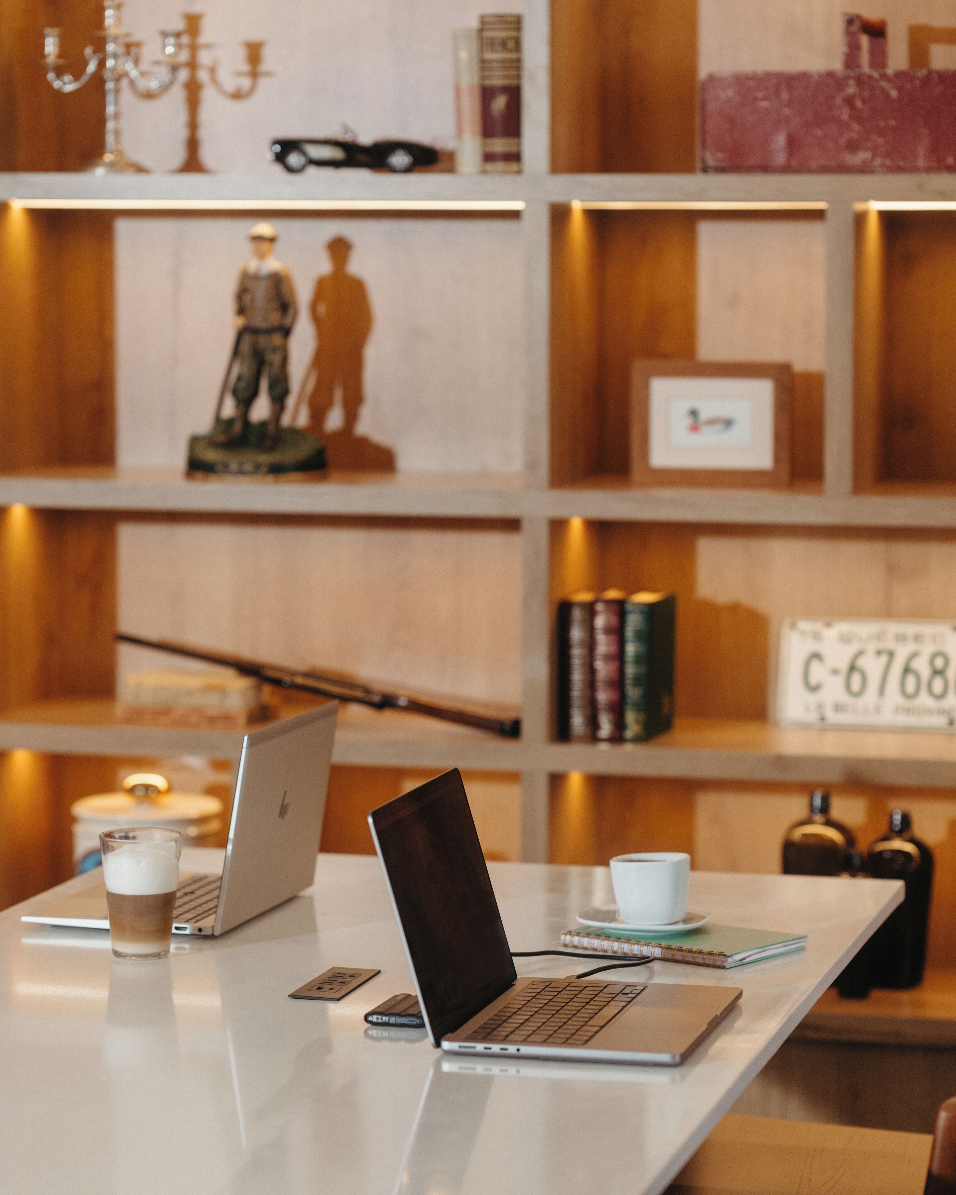 Un espacio de trabajo con dos portátiles, una taza de café, un cuaderno y una taza. Estantes al fondo exhiben libros, objetos decorativos y un cuadro enmarcado.