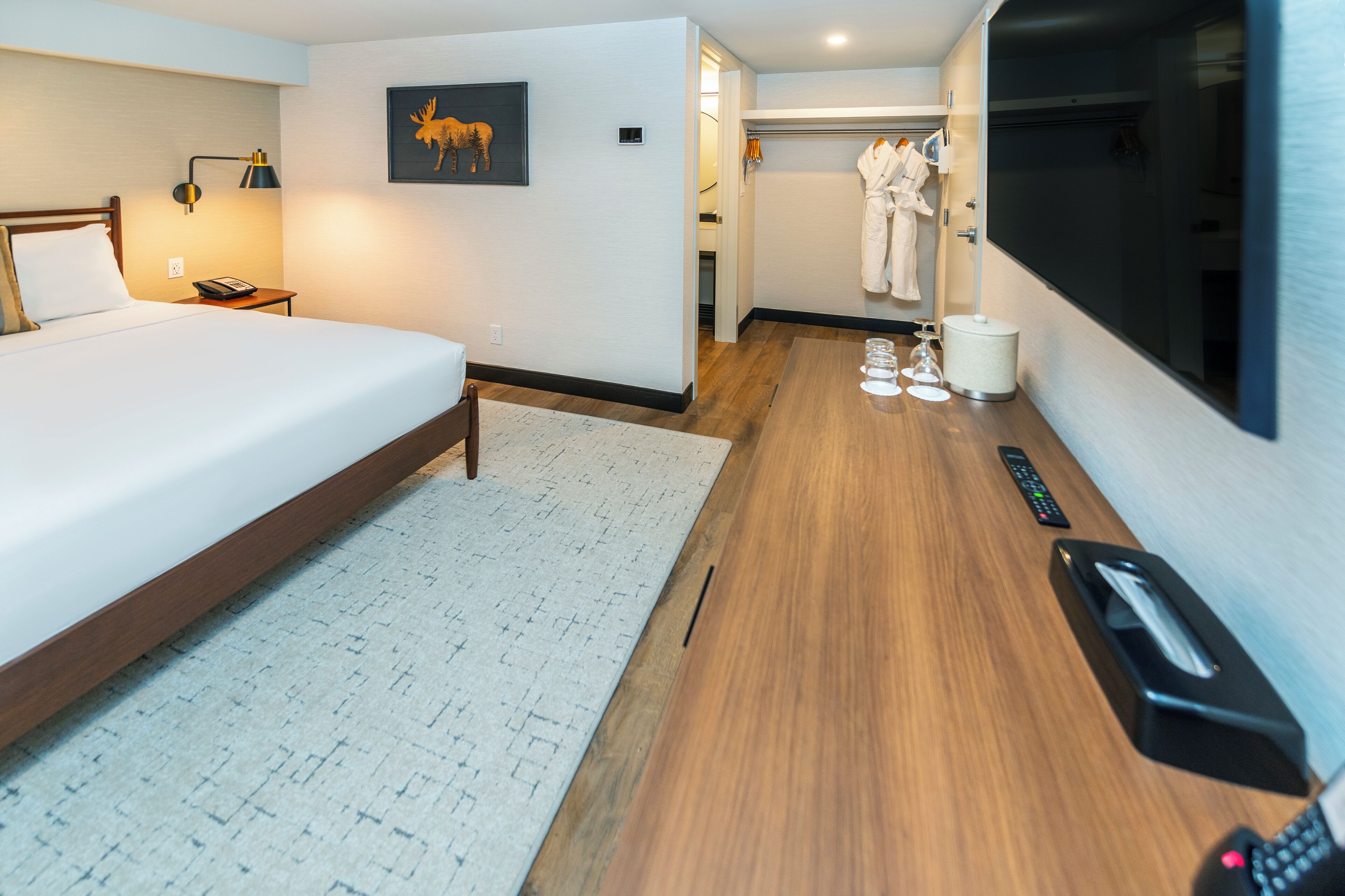 A hotel room with a large bed, a bedside table with a lamp, a wall-mounted TV, and a small bathroom with a shower and a towel rack.