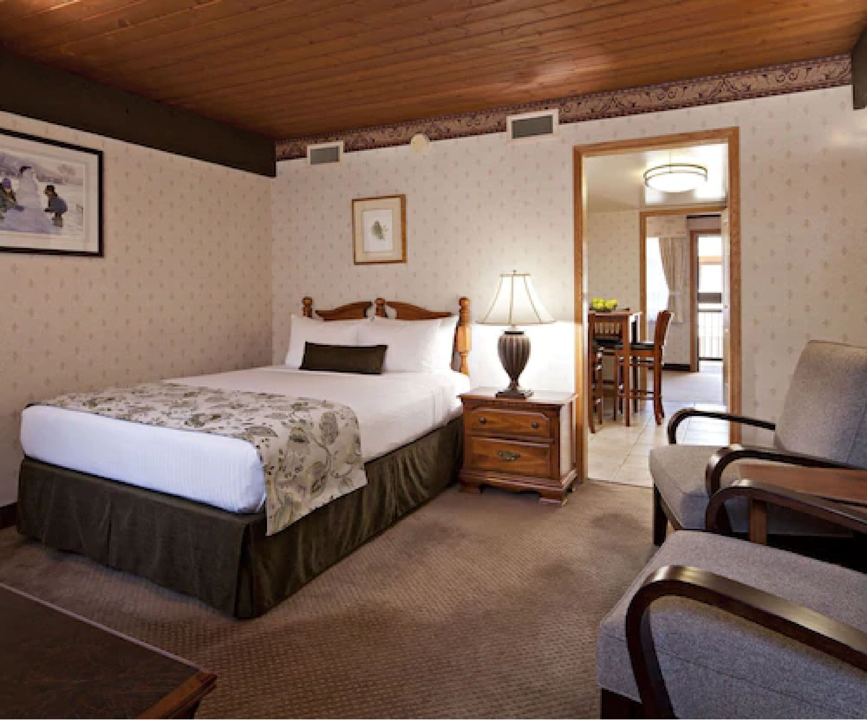 Una habitación de hotel con una cama grande, una mesita de noche de madera, un escritorio con una lámpara, una silla y un área de comedor con una mesa y sillas.