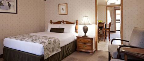 A hotel room with a large bed, a wooden nightstand, a desk with a lamp, a chair, and a dining area with a table and chairs.